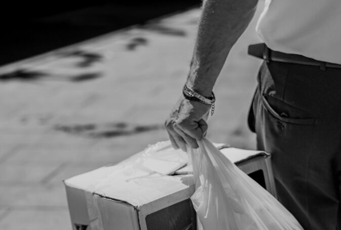 A black and white photo of a man carrying a box and plastic bag on a city street, showcasing urban daily life.